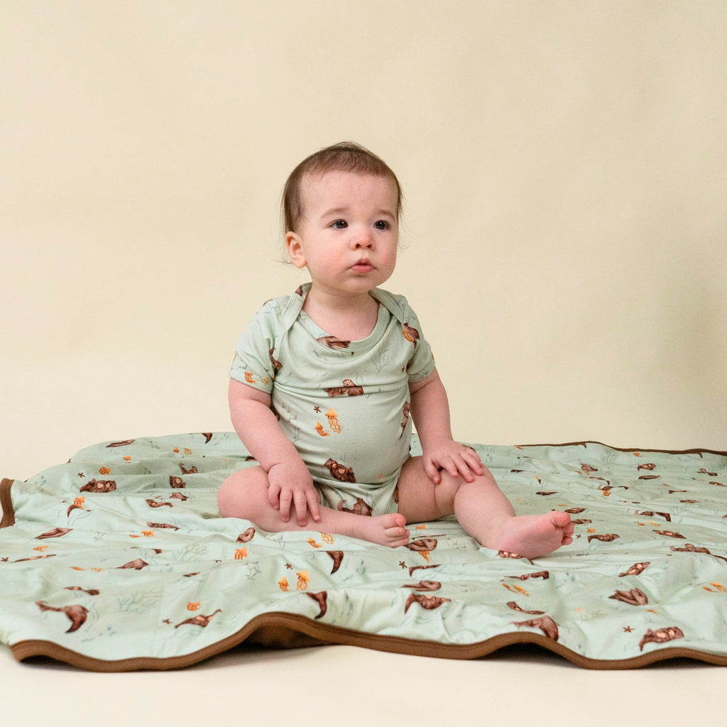 baby wearing onesie in otters print, sitting on quilted otters child blanket