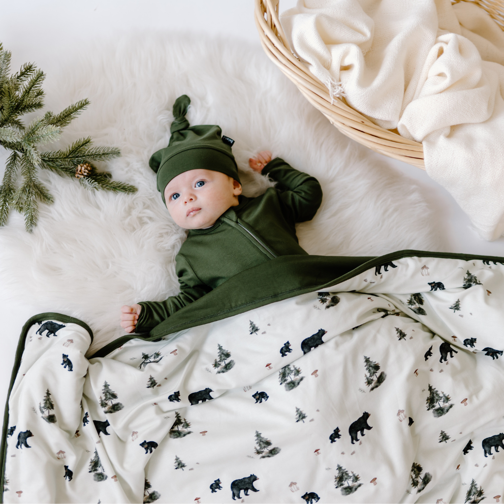 baby wearing forest green hat and sleeper and covered until belly with child blanket in black bears print